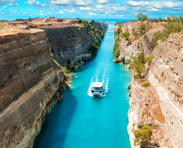 Beautiful scenery of the Corinth Canal in a bright sunny day against a blue sky with white clouds. Among the rocks floating white ship in turquoise water.